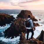 man standing near cliff looking at body of water during daytime